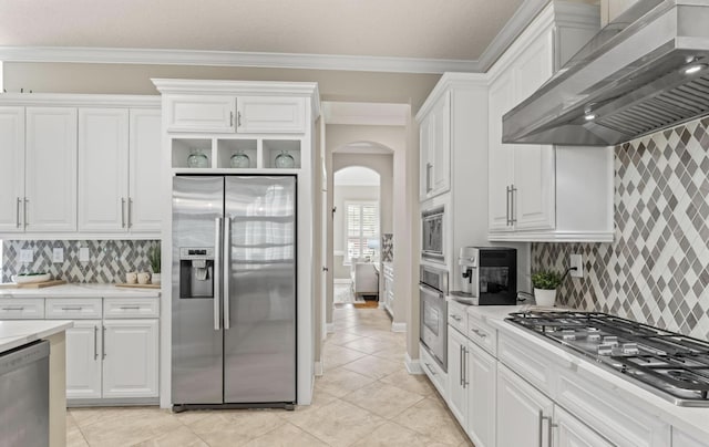 kitchen with wall chimney exhaust hood, stainless steel appliances, backsplash, crown molding, and white cabinets