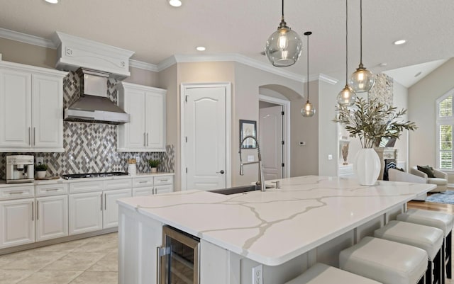 kitchen featuring white cabinets, a center island with sink, hanging light fixtures, light stone countertops, and beverage cooler