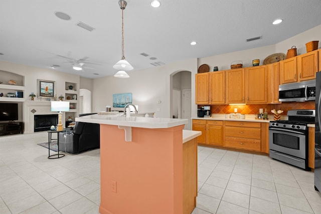 kitchen with light tile patterned floors, built in features, appliances with stainless steel finishes, a center island with sink, and decorative light fixtures