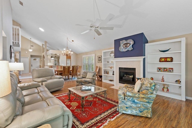 living room with built in features, vaulted ceiling, wood finished floors, a tile fireplace, and ceiling fan with notable chandelier