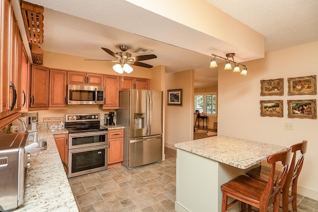 kitchen featuring ceiling fan, appliances with stainless steel finishes, brown cabinets, light stone counters, and a kitchen breakfast bar