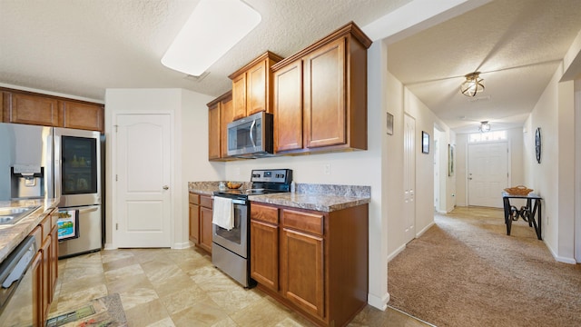 kitchen featuring appliances with stainless steel finishes, brown cabinetry, a textured ceiling, and light colored carpet