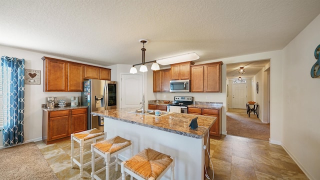 kitchen featuring a breakfast bar area, a kitchen island with sink, stainless steel appliances, hanging light fixtures, and brown cabinets