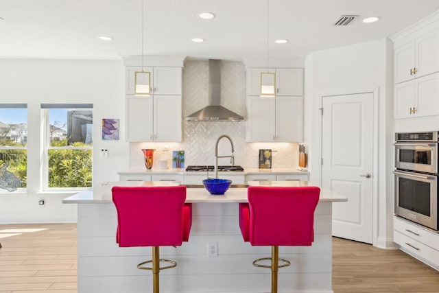 kitchen featuring a kitchen island with sink, light countertops, appliances with stainless steel finishes, wall chimney range hood, and tasteful backsplash