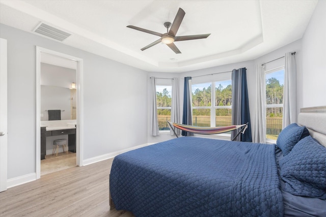 bedroom featuring ceiling fan, ensuite bath, a tray ceiling, and light hardwood / wood-style floors