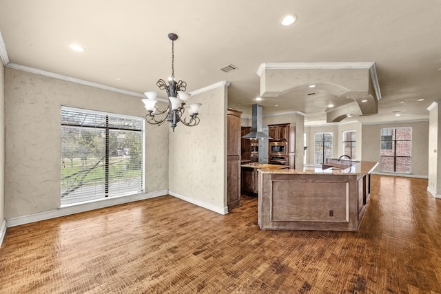 kitchen featuring light stone countertops, a notable chandelier, an island with sink, wall chimney exhaust hood, and pendant lighting