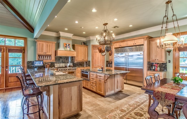 kitchen with high end appliances, backsplash, custom range hood, dark stone countertops, and an inviting chandelier