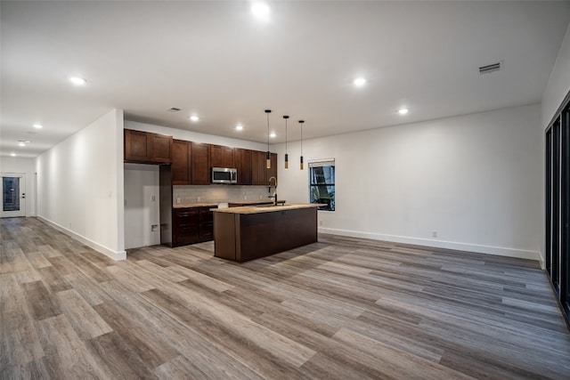 kitchen with pendant lighting, stainless steel appliances, light wood-type flooring, a kitchen island with sink, and backsplash