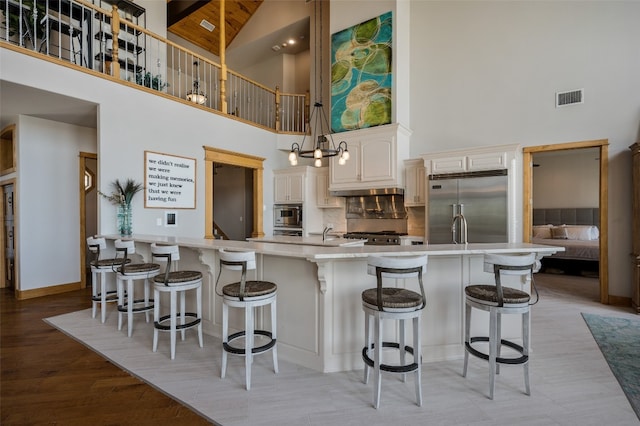 kitchen with light hardwood / wood-style floors, a kitchen breakfast bar, built in appliances, high vaulted ceiling, and a kitchen island with sink