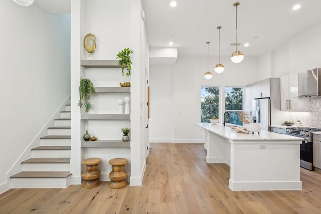 kitchen featuring wall chimney exhaust hood, light hardwood / wood-style floors, pendant lighting, a kitchen island with sink, and appliances with stainless steel finishes