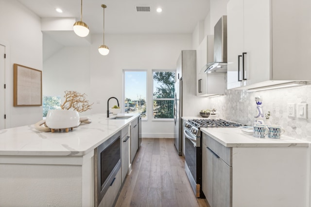 kitchen featuring wall chimney exhaust hood, pendant lighting, stainless steel appliances, light stone countertops, and a kitchen island with sink