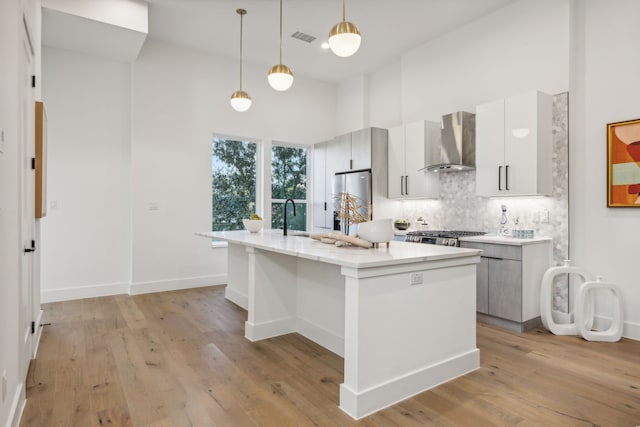 kitchen featuring a kitchen island with sink, wall chimney range hood, decorative light fixtures, white cabinets, and light hardwood / wood-style floors