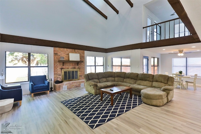 living room featuring ceiling fan, beamed ceiling, a brick fireplace, light wood-type flooring, and a towering ceiling