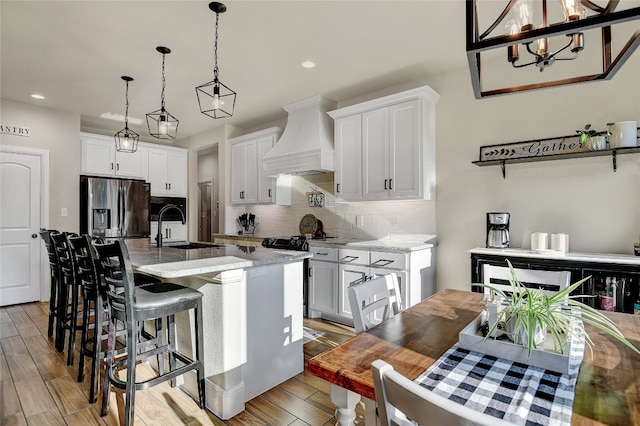 kitchen with an island with sink, stainless steel fridge, pendant lighting, custom exhaust hood, and white cabinetry