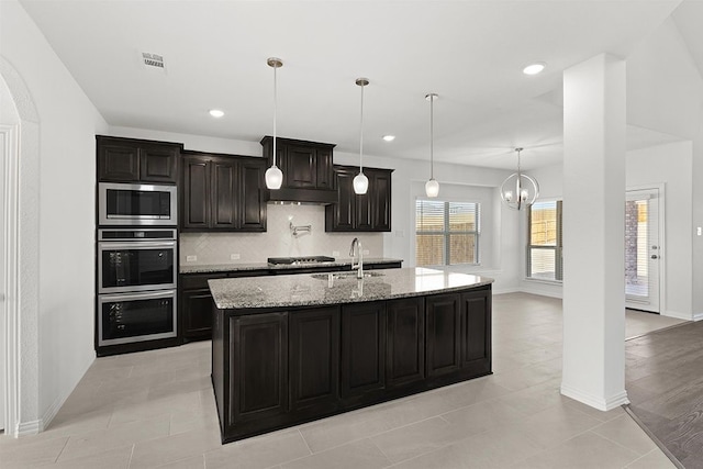 kitchen featuring light stone counters, backsplash, decorative light fixtures, stainless steel appliances, and a center island with sink