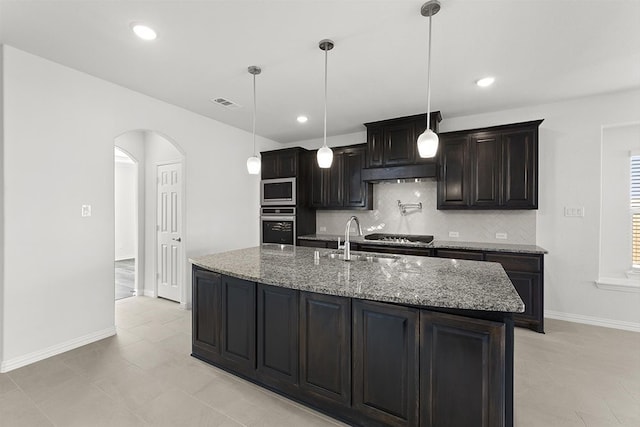 kitchen with appliances with stainless steel finishes, hanging light fixtures, light tile flooring, dark stone counters, and a kitchen island with sink