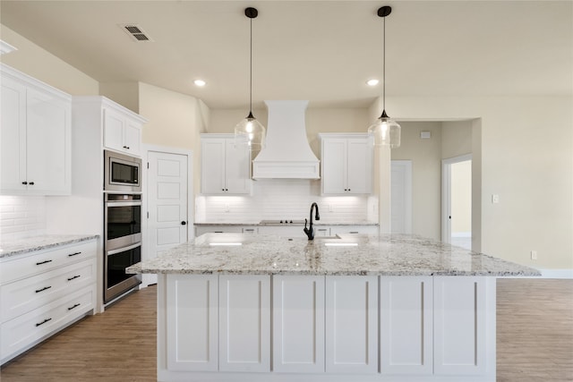 kitchen featuring light wood-type flooring, premium range hood, pendant lighting, a center island with sink, and white cabinetry