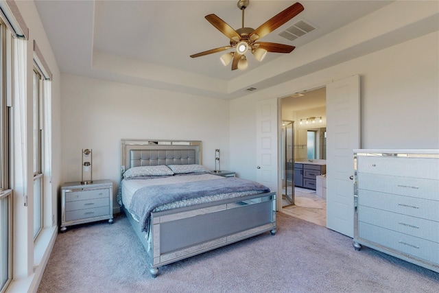 bedroom with ceiling fan, light colored carpet, a raised ceiling, and ensuite bath