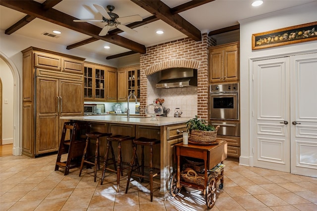 kitchen with coffered ceiling, beam ceiling, wall chimney range hood, ceiling fan, and a kitchen island with sink