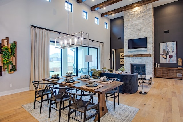 dining space with light hardwood / wood-style floors, a notable chandelier, beamed ceiling, a stone fireplace, and a towering ceiling