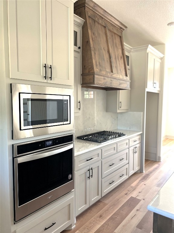 kitchen with custom exhaust hood, stainless steel appliances, decorative backsplash, white cabinets, and light hardwood / wood-style floors