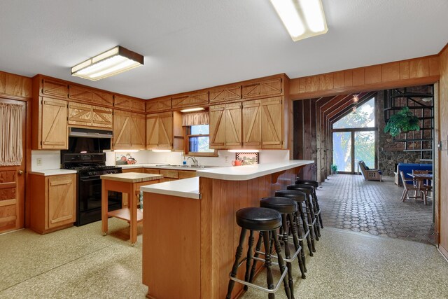 kitchen featuring black gas range, sink, kitchen peninsula, wood walls, and a breakfast bar area