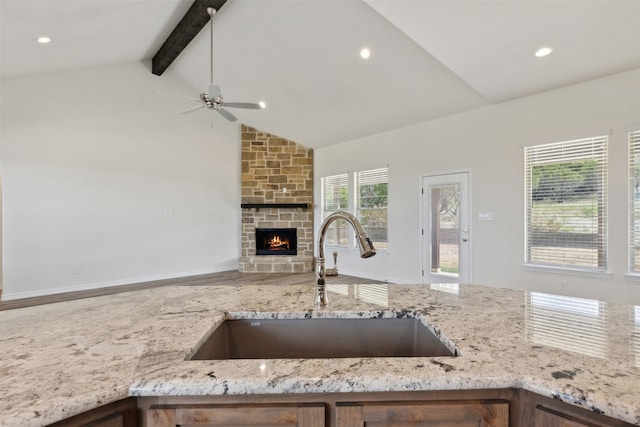 kitchen featuring ceiling fan, light stone countertops, vaulted ceiling with beams, a stone fireplace, and sink