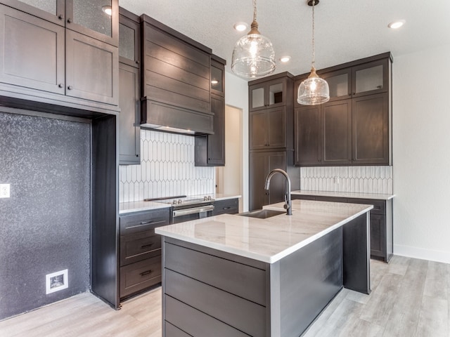 kitchen with custom range hood, an island with sink, sink, and light wood-type flooring