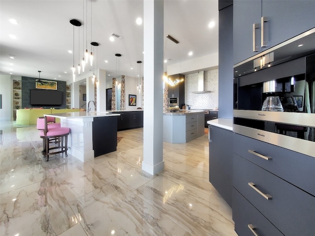 kitchen with an island with sink, decorative light fixtures, wall chimney range hood, a kitchen bar, and backsplash