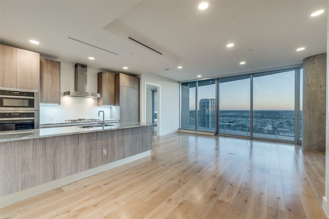 kitchen featuring double oven, sink, light wood-type flooring, floor to ceiling windows, and wall chimney range hood