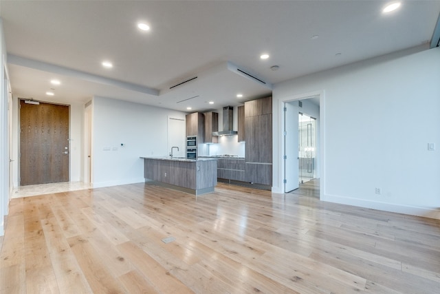 kitchen with wall chimney range hood, a breakfast bar area, a kitchen island with sink, and light hardwood / wood-style flooring