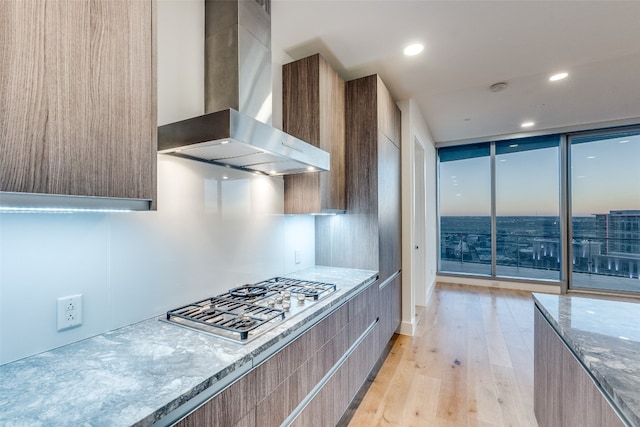 kitchen with wall chimney exhaust hood, stainless steel gas cooktop, light stone counters, and light hardwood / wood-style flooring