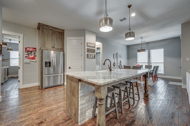 kitchen featuring an island with sink, hanging light fixtures, a breakfast bar, stainless steel fridge, and light stone counters