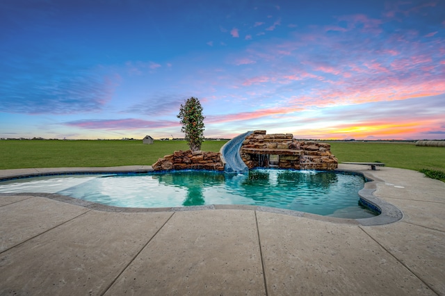 pool at dusk featuring pool water feature, a lawn, and a water slide