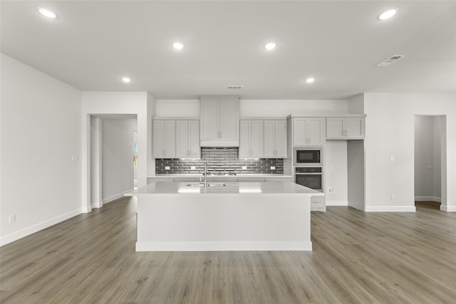 kitchen with wood-type flooring, tasteful backsplash, black microwave, an island with sink, and oven