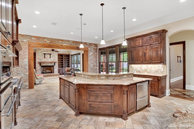 kitchen with pendant lighting, a fireplace, light stone countertops, backsplash, and a kitchen island with sink