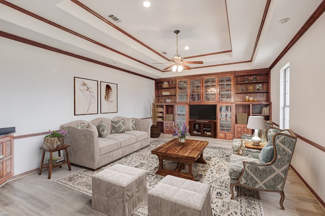 living room featuring light hardwood / wood-style flooring, built in shelves, ceiling fan, a raised ceiling, and ornamental molding