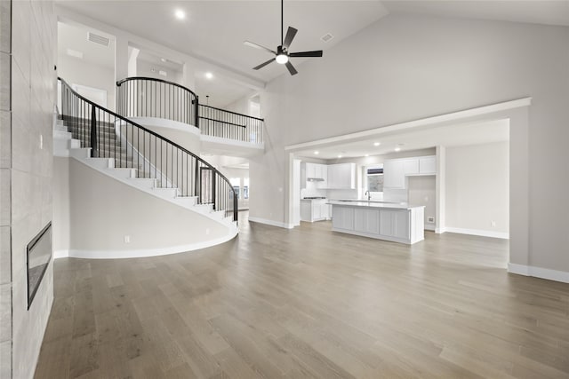 unfurnished living room with sink, high vaulted ceiling, ceiling fan, a tiled fireplace, and light hardwood / wood-style floors