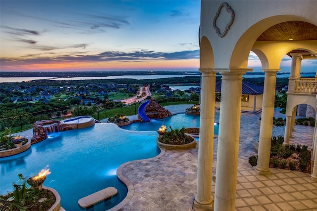 pool at dusk with a patio, pool water feature, a water view, and an in ground hot tub