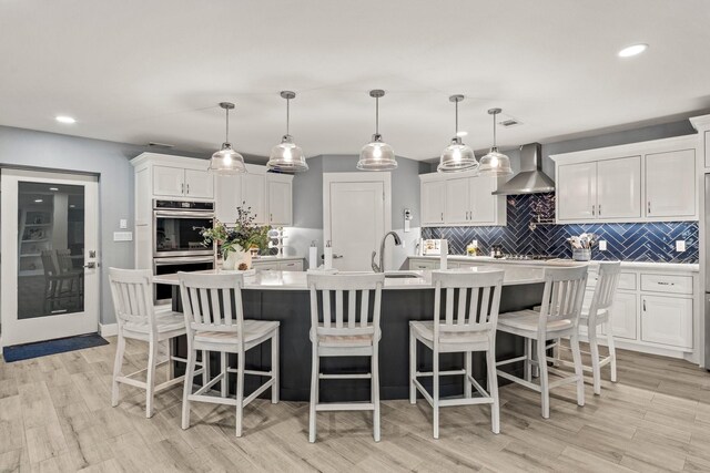 kitchen with tasteful backsplash, a kitchen island with sink, white cabinetry, wall chimney exhaust hood, and decorative light fixtures