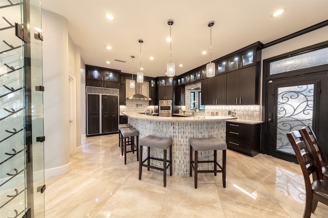 kitchen featuring dark brown cabinetry, stainless steel oven, a center island with sink, hanging light fixtures, and wall chimney range hood