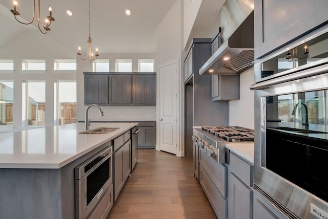 kitchen featuring appliances with stainless steel finishes, sink, wall chimney range hood, a chandelier, and an island with sink