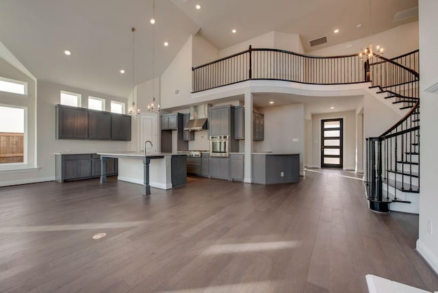 interior space with pendant lighting, a towering ceiling, a center island with sink, and wall chimney range hood