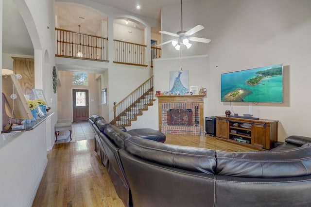 living room with wood-type flooring, ceiling fan, a towering ceiling, and a brick fireplace