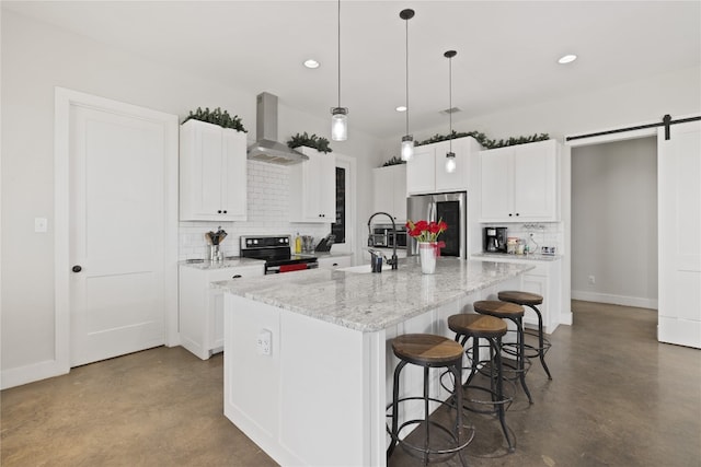 kitchen featuring a kitchen island with sink, wall chimney range hood, a barn door, appliances with stainless steel finishes, and concrete floors