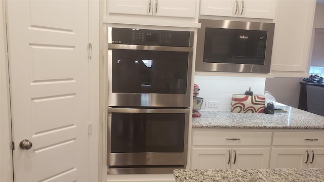 kitchen with built in microwave, backsplash, white cabinets, light stone counters, and stainless steel double oven