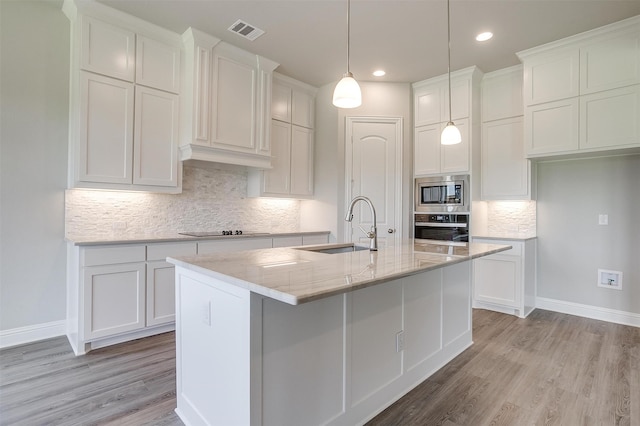 kitchen with appliances with stainless steel finishes, light wood-type flooring, a kitchen island with sink, and backsplash