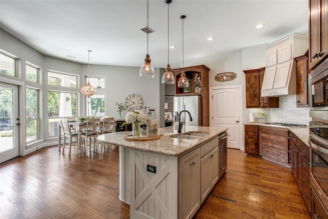 kitchen with appliances with stainless steel finishes, hanging light fixtures, a kitchen island with sink, sink, and backsplash