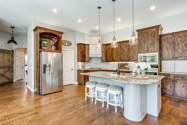 kitchen featuring appliances with stainless steel finishes, tasteful backsplash, hanging light fixtures, light stone counters, and a center island with sink