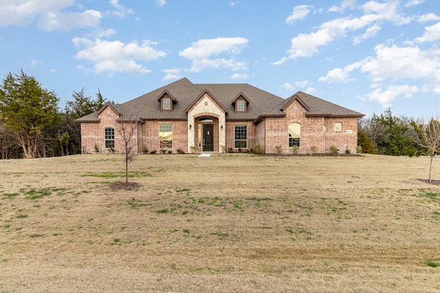 french country home featuring roof with shingles, brick siding, and a front lawn